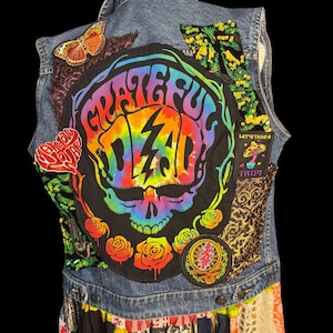 Könnte beinhalten: Eine blaue Jeansweste mit einem großen Grateful Dead Totenkopf-Aufnäher in Regenbogenfarben, umgeben von kleineren Aufnähern wie einem Schmetterling, einem Pilz mit dem Text Let's Take A Trip und grünen Pailletten auf dunklem Stoff.