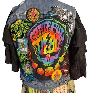 May include: A denim jacket with a variety of patches and embellishments. The back features a large, colorful "Grateful Dead" skull design, surrounded by roses. Additional patches include a sun, a lightning bolt, and other symbols. The sleeves are black with ruffles.