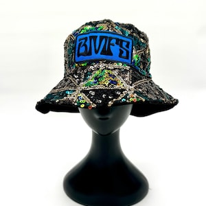 Peut inclure: Un chapeau bob recouvert de sequins noirs, verts et dorés disposés en grille géométrique. L'avant présente un écusson bleu rectangulaire avec le texte noir BMFS brodé dans une police stylisée. Le chapeau possède un bord large et incliné.