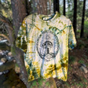 May include: A short-sleeve t-shirt featuring a tie-dye pattern in shades of green, yellow, and white. The center graphic is an oval frame containing a mushroom, a stone path, and the text "SPRING TOUR" and "2025" in a black ink illustration.