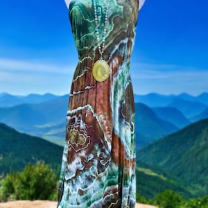Op de afbeelding: Een lange, mouwloze maxi-jurk met een tie-dye-patroon in tinten groen, bruin en wit. De jurk heeft een losse pasvorm en een vloeiende silhouet. Het is versierd met een lange ketting met een grote, ronde hanger.