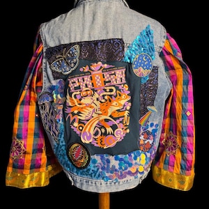 May include: A denim jacket with colorful patches and sequin embellishments. The back features a large graphic with text, a butterfly, and other decorative elements. The sleeves are a vibrant plaid pattern with gold trim.