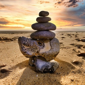 Puede incluir: Una pila de piedras grises equilibradas sobre una piedra gris más grande e irregular, con el telón de fondo de una playa de arena y un cielo de puesta de sol vibrante.