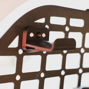 Puede incluir: Un gancho de metal negro montado en un panel de rejilla de metal negro. El gancho está fijado con dos tornillos. El panel de rejilla tiene un patrón cuadrado.