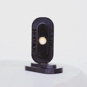 Puede incluir: Un objeto negro impreso en 3D con el texto "MOJAVE MFG" en el lateral. El objeto tiene forma ovalada con un agujero circular y se apoya sobre una base rectangular. El fondo es una superficie blanca lisa.