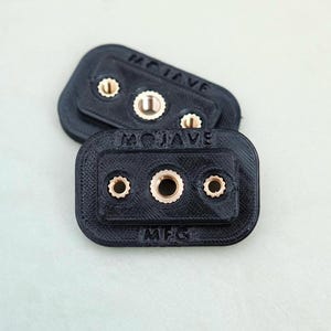 Puede incluir: Dos placas rectangulares de plástico negro con el texto "MOJAVE MFG" impreso en ellas. Cada placa tiene tres insertos de metal de color dorado.