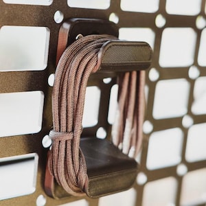 Könnte beinhalten: Ein braunes Kabel ist um einen schwarzen, aus Kunststoff gefertigten Kabelorganisator gewickelt. Der Organisator ist an einer Metallgitterwand montiert.