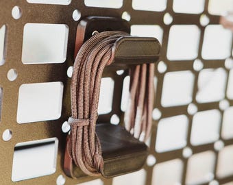 May include: A brown cord is wrapped around a black plastic cord organizer. The organizer is mounted on a metal grid wall.
