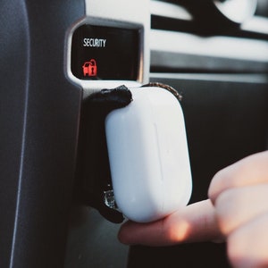 Può includere: Una custodia di ricarica wireless bianca con un cinturino nero attaccato al cruscotto di un'auto. La custodia è tenuta da una mano. La spia di sicurezza dell'auto è accesa e dice "SECURITY".