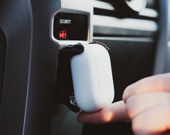 Puede incluir: Una funda de carga inalámbrica blanca con una correa negra unida al tablero de un automóvil. La funda está siendo sostenida por una mano. La luz de seguridad del automóvil está encendida y dice "SECURITY".