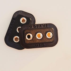 Puede incluir: Tres objetos pequeños, rectangulares y oscuros con ojales metálicos. Los dos superiores son negros y el inferior es marrón. La palabra "MOJAVE" está grabada en los objetos negros y "MOJAVE MFG" en el objeto marrón.