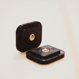 Puede incluir: Dos accesorios de cámara negros, de forma cuadrada, con un inserto roscado de color dorado. Uno está en posición vertical y el otro está plano. Probablemente se utilizan para montar cámaras u otros equipos.