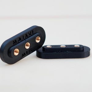 Puede incluir: Dos bloques de conexión de plástico negro con terminales de tornillo de color dorado. El bloque más grande tiene el texto "MOJAVE MFG" impreso en él.