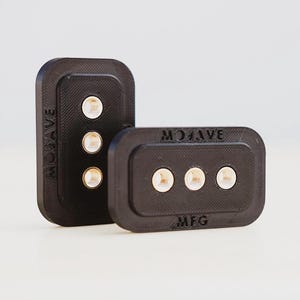 Puede incluir: Dos bloques rectangulares de plástico negro con tres pequeños círculos blancos en cada uno. Los bloques tienen el texto "MOGAVE" y "MFG" en relieve.