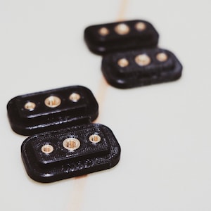 Puede incluir: Dos bloques rectangulares de plástico negro con tres tornillos dorados cada uno. Los bloques están apilados uno encima del otro.