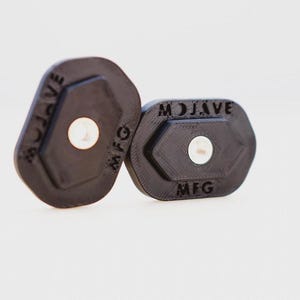 Puede incluir: Dos objetos hexagonales de plástico negro con el texto "MOJAVE MFG" impreso en ellos.