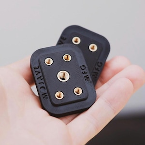 Puede incluir: Dos placas de montaje de cámara de plástico negro con tornillos de color dorado y el texto "WAVE MFG" impreso en ellas.