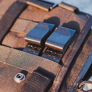 May include: Close-up of a brown tactical bag with two black MOLLE clips attached. The clips are labeled "MOLLE MFG".