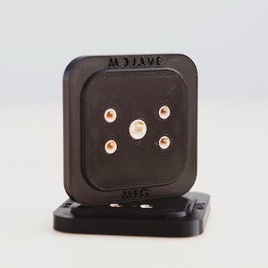 Puede incluir: Dispositivo cuadrado negro con esquinas redondeadas, con el texto "MOJAVE" y "MFG". El dispositivo tiene varios elementos circulares dorados y un componente central plateado. Descansa sobre una base negra a juego.