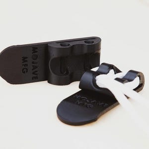 May include: Two black cord organisers with the text "MOJAVE MFG" embossed on them. One organiser holds white cords. The organisers are made of a matte black material and are designed to manage cords.