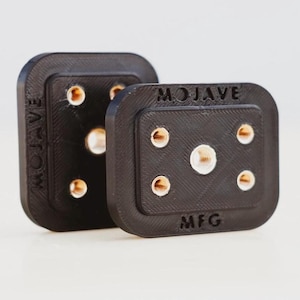 Puede incluir: Dos cuadrados de plástico negro con el texto "MOJAVE MFG" en uno de ellos. Cada cuadrado tiene seis pequeños agujeros redondos de color dorado.