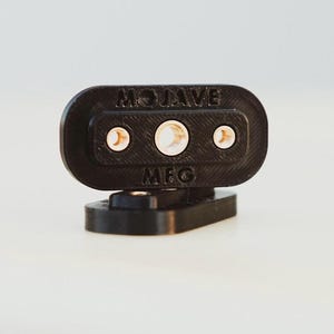 Puede incluir: Soporte de montaje de plástico negro con tres agujeros y el texto "MOJAVE MFG" impreso en él.