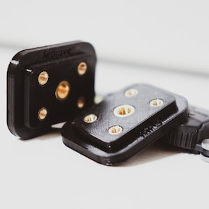May include: Three black plastic camera mounts with gold-coloured screws. The mounts have a square shape and are designed to attach to a camera tripod.