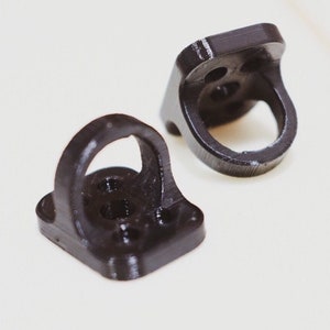 Puede incluir: Dos soportes de plástico negro impresos en 3D con un agujero circular en el centro. Los soportes tienen una base cuadrada con cuatro agujeros.
