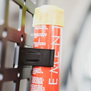 Op de afbeelding: Een rode en gele fakkel met een zwarte plastic clip bevestigd aan een metalen rooster. De fakkel heeft het woord "MINUT" op de zijkant gedrukt.