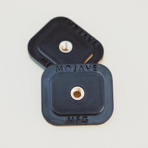 Puede incluir: Dos soportes de cámara cuadrados de plástico negro con un agujero roscado en el centro. Los soportes están etiquetados con "MOJAVE MFG".