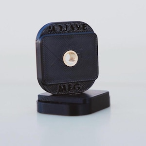 Puede incluir: Un soporte de cámara de plástico negro con el texto "MOJAVE MEG" impreso en él. El soporte tiene un agujero roscado en el centro.