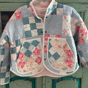May include: A quilted jacket in a patchwork design, featuring squares of blue, pink, and white fabrics. The jacket has a round collar, two pockets, and a button closure. The lining is a pink gingham pattern.