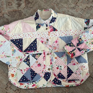 May include: A quilted jacket featuring a patchwork design of various floral, gingham, and polka dot patterns in shades of pink, navy blue, red, and cream. The jacket has a high collar, long sleeves, and two large front patch pockets.