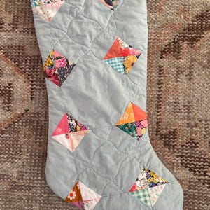 May include: A quilted Christmas stocking in light blue fabric featuring a repeating pattern of diamond-shaped patches. Each diamond is composed of four smaller triangles in various floral, gingham, and solid colors including pink, orange, and green.