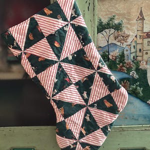 May include: A quilted Christmas stocking featuring a geometric pattern of alternating dark green fabric with a dog print and white fabric with thin red stripes. The stocking is constructed from triangular fabric segments stitched together.