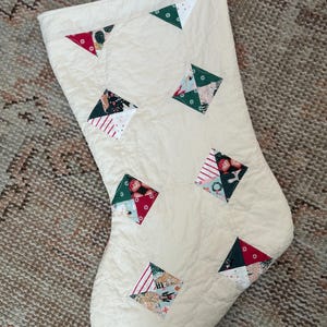 May include: A quilted Christmas stocking in an off-white fabric featuring a repeating pattern of diamond-shaped patches. Each diamond is composed of smaller triangular segments in festive red, dark green, and white with various holiday-themed prints.