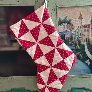 May include: A quilted Christmas stocking featuring a geometric pinwheel pattern. The design consists of alternating triangles of solid off-white fabric and a vibrant red fabric decorated with a small, repeating white floral print throughout.