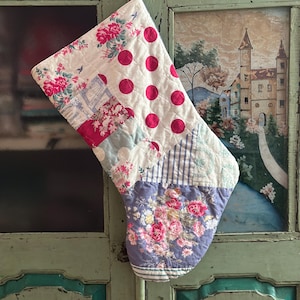 May include: A quilted Christmas stocking featuring a patchwork design of various fabrics. The patterns include bright pink polka dots on white, a floral print with pink roses on a white background, and a blue and white floral print on the toe area.