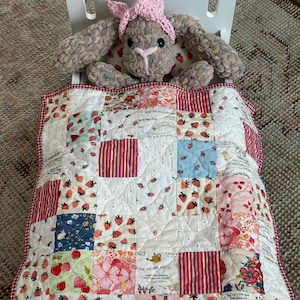 May include: A plush gray rabbit with a pink crocheted headband resting in a white wooden doll bed. The rabbit is covered by a patchwork quilt featuring strawberry prints, floral patterns, and red stripes, finished with a red and white gingham trim.