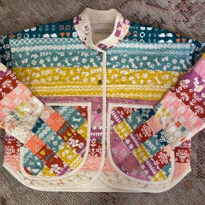 May include: A quilted jacket featuring horizontal stripes of various floral and geometric patterns in teal, yellow, pink, and red. The garment has a stand-up collar, a front button closure, and two large patch pockets with a colorful patchwork design.