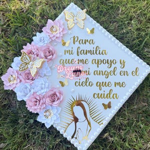 May include: A white graduation cap decorated with pink and white paper flowers, gold butterflies, and a gold image of the Virgin Mary. The cap has the text "Para mi familia que me apoyo y mi angel en el cielo que me cuida" in gold lettering.