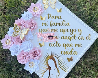 May include: A white graduation cap decorated with pink and white paper flowers, gold butterflies, and a gold image of the Virgin Mary. The cap has the text "Para mi familia que me apoyo y mi angel en el cielo que me cuida" in gold lettering.
