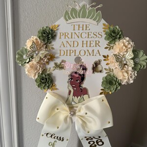 May include: A graduation cap decorated with a princess theme. The cap features the text "THE PRINCESS AND HER DIPLOMA" in gold lettering, along with a cartoon image of a princess. The cap is adorned with cream and green paper flowers, butterflies, and a large cream bow.