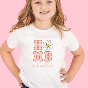 K&ouml;nnte beinhalten: Ein wei&szlig;es T-Shirt mit einer pinkfarbenen und orangenen Grafik, auf der "HOME" mit einer G&auml;nsebl&uuml;mchen und "schooled" darunter steht.