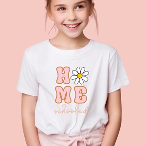 K&ouml;nnte beinhalten: Ein wei&szlig;es T-Shirt mit einer pinkfarbenen und orangenen Grafik, auf der "HOME" mit einer G&auml;nsebl&uuml;mchen im "O" und "schooled" darunter steht.