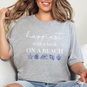 May include: A heather gray short-sleeve t-shirt featuring the white text "happiest with a book ON A BEACH" above a row of blue graphic icons including a starfish, a nautilus shell, a wave, a conch shell, and a piece of coral.