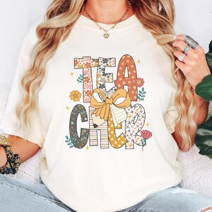May include: Cream-colored t-shirt with the word "TEACHER" in a colorful, floral design. The letters are decorated with flowers, polka dots, and a bow. A pencil and notebook are also featured in the design.