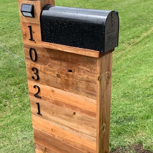 May include: A black metal mailbox mounted on a vertical wooden post structure. The post features horizontal wood planks and displays the house numbers 10321 in black metal digits. A small solar light is attached to the top left of the wooden post.