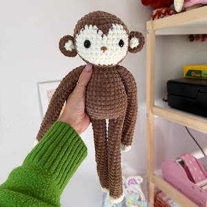 May include: A handmade crochet monkey doll in shades of brown and cream. The monkey has a detailed face with black eyes and a tan nose. The arms and legs are long and slender, with cream-colored accents on the hands and feet. The doll is soft and cuddly.