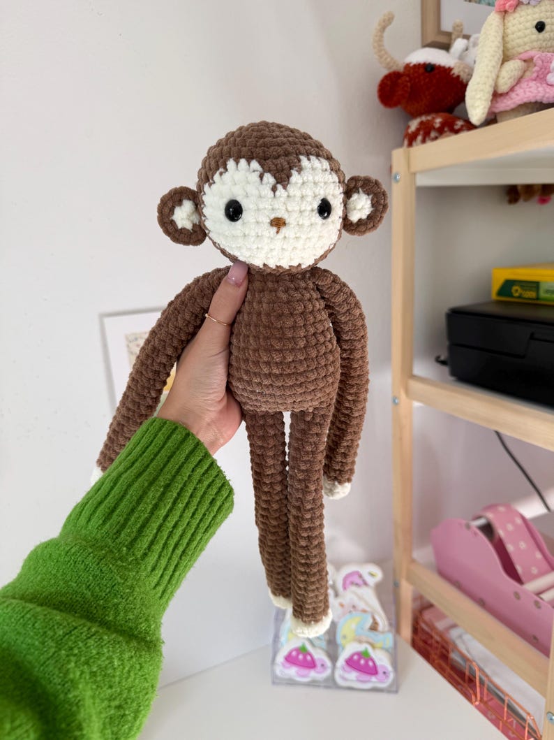 May include: A handmade crochet monkey doll in shades of brown and cream. The monkey has a detailed face with black eyes and a tan nose. The arms and legs are long and slender, with cream-colored accents on the hands and feet. The doll is soft and cuddly.